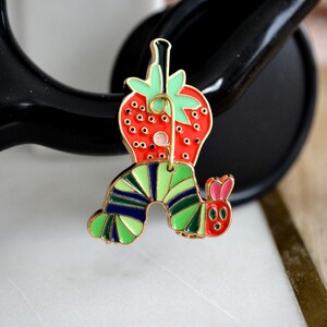 May include: A colorful enamel pin featuring a strawberry and a caterpillar design. The strawberry is red with green leaves, and the caterpillar has green, blue, and yellow stripes. The pin has a gold-colored outline.