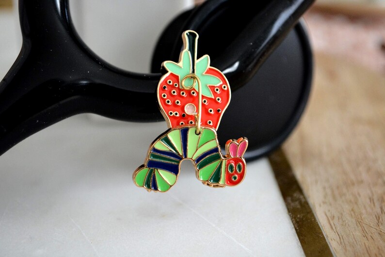 May include: A colorful enamel pin featuring a strawberry and a caterpillar design. The strawberry is red with green leaves, and the caterpillar has green, blue, and yellow stripes. The pin has a gold-colored outline.