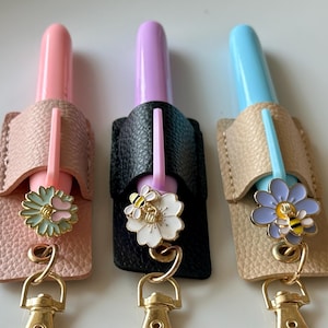 May include: Three hand sanitiser holders in pink, purple, and blue, each with a different coloured sanitiser inside. The holders are made of textured leather and have gold-coloured hardware. Each features a flower charm with a bee.