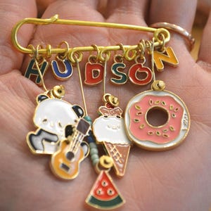 May include: A gold safety pin brooch with the name "HUDSON" in colourful letters. Charms include a panda playing a guitar, an ice cream cone, a watermelon slice, and a pink donut.