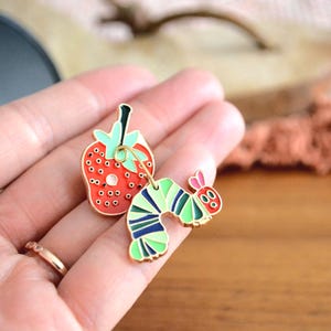 May include: A pair of colorful enamel pins featuring a red strawberry and a green, blue, and yellow caterpillar. The strawberry has black seeds and a green stem. The caterpillar has pink antennae and a smiling face.