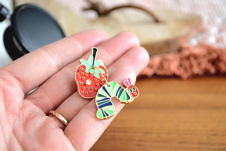 May include: A pair of colorful enamel pins featuring a red strawberry and a green, blue, and yellow caterpillar. The strawberry has black seeds and a green stem. The caterpillar has pink antennae and a smiling face.