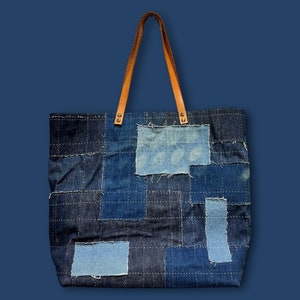 Może przedstawiać: Dżinsowa torba typu tote z patchworkowym wzorem w różnych odcieniach niebieskiego. Torba posiada jasnobrązowe skórzane uchwyty przymocowane metalowymi nitami. Torba wykonana jest z dżinsu i ma prostokątny kształt.