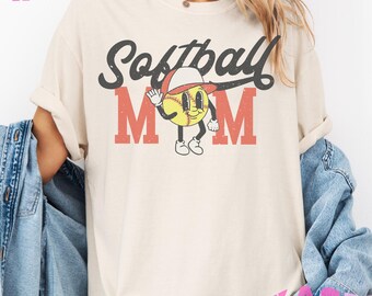 May include: A cream colored t-shirt with the text "Softball Mom" in black and red. A cartoon softball with a face and a baseball cap is in the center of the text.