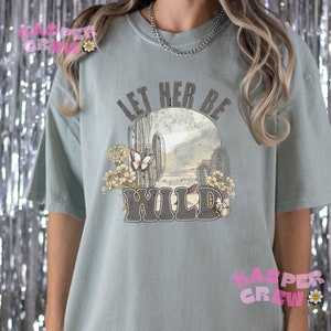 May include: A light blue t-shirt with a graphic print of a cactus and butterflies. The text "Let Her Be Wild" is printed on the shirt.