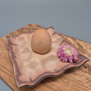Può includere: Un uovo marrone con un cuore disegnato su di esso si trova in un porta uova in ceramica rosa e verde. Un fiore rosa è accanto all'uovo.