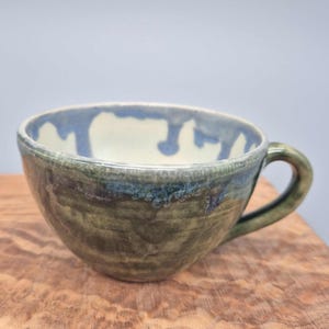 Könnte beinhalten: Handgefertigte Keramiktasse mit grüner und blauer Glasur. Die Tasse hat eine runde Form mit Henkel und hellem Innenraum. Die Außenseite weist eine Mischung aus grünen und blauen Farbtönen auf, die einen einzigartigen, künstlerischen Look erzeugt. Die Tasse steht auf einer Holzoberfläche.