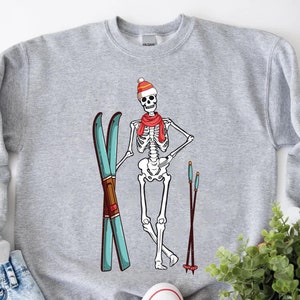 May include: A grey sweatshirt featuring a skeleton wearing a red and white hat and a red scarf, holding blue and white skis. The skeleton is standing against a white background.