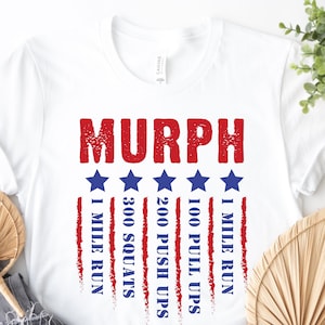 May include: White t-shirt with the word "MURPH" in red letters and a red, white, and blue American flag design. The flag design includes the words "1 mile run", "100 pull ups", "200 push ups", and "300 squats".