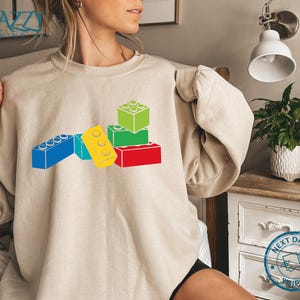 May include: A beige sweatshirt featuring a colorful graphic of toy building blocks in red, yellow, blue, and green. The blocks are stacked and arranged to create a playful design. The sweatshirt has a crew neck and long sleeves.