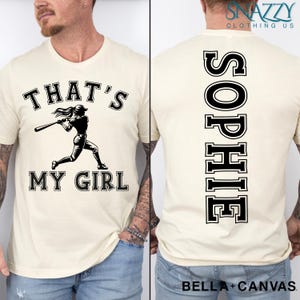 May include: A white t-shirt with a black graphic of a baseball player and the text "That's My Girl". The back of the shirt has the name "Sophie" printed vertically in black.