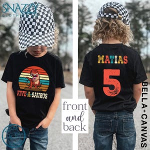 May include: Black t-shirt featuring a vintage-style illustration of a dinosaur with the text "Five-A-saurus" on the front. The back of the shirt has the name "Matias" and the number "5" in colourful, distressed lettering.