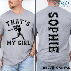 May include: Gray t-shirt with a baseball player graphic and the text "That's My Girl". The back of the shirt has the name "Sophie" printed vertically in a varsity style font.