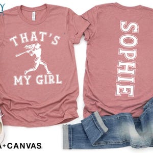 May include: A pink t-shirt with the text "That's My Girl" and a graphic of a baseball player on the front. The back of the shirt has the name "Sophie" printed vertically down the back.