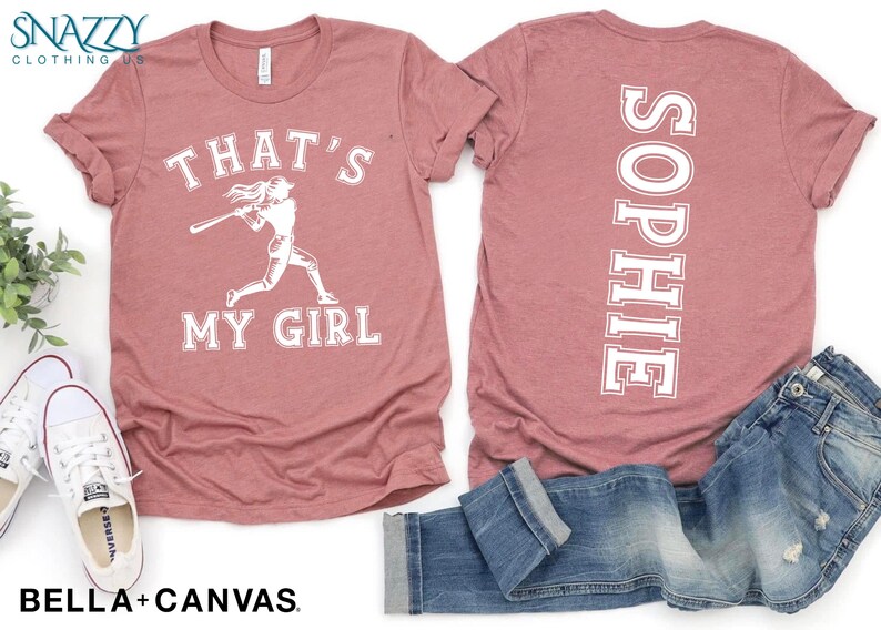 May include: A pink t-shirt with the text "That's My Girl" and a graphic of a baseball player on the front. The back of the shirt has the name "Sophie" printed vertically down the back.