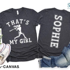 May include: Two dark gray t-shirts with white text. One shirt says "That's My Girl" with a graphic of a girl playing baseball. The other shirt has the name "Sophie" printed vertically down the back.