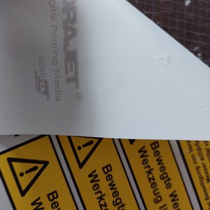 May include: A close-up of a white surface with the text "ORAJET Digital Printing Media" and the ORAFOL logo. Below, a yellow warning label with black text and a warning symbol is partially visible.