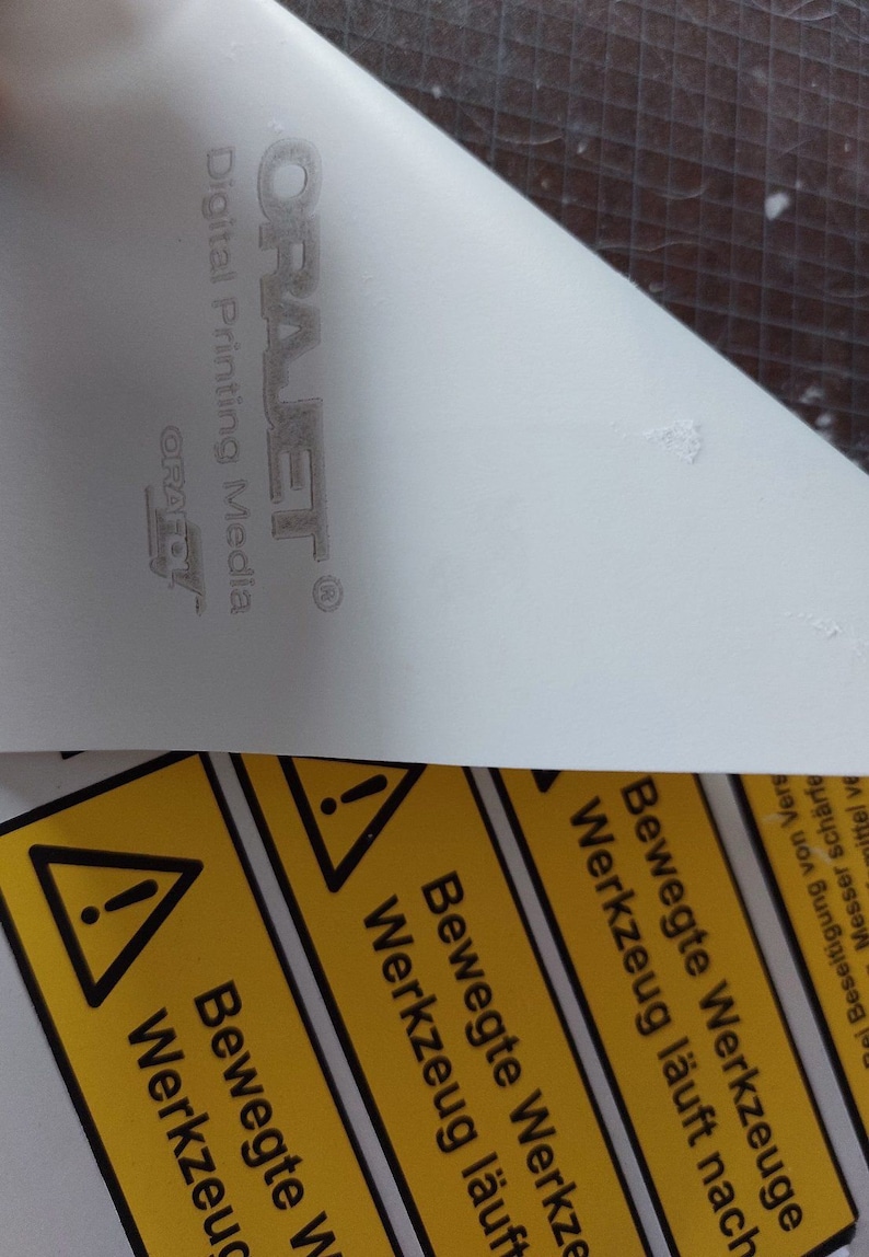 May include: A close-up of a white surface with the text "ORAJET Digital Printing Media" and the ORAFOL logo. Below, a yellow warning label with black text and a warning symbol is partially visible.