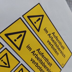 May include: Yellow rectangular warning labels with a black triangle and exclamation point symbol. The text "Aufenthalt im Arbeitsbereich verboten" is printed in black. The labels are on a white surface.