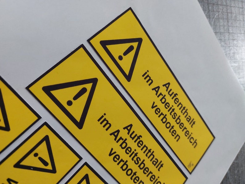 May include: Yellow rectangular warning labels with a black triangle and exclamation point symbol. The text "Aufenthalt im Arbeitsbereich verboten" is printed in black. The labels are on a white surface.