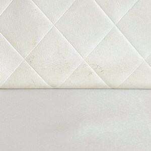 May include: Close-up of a white, quilted leather surface with a diamond pattern. The leather is slightly folded over, revealing a smooth, white surface below. Some areas of the leather show minor discoloration.