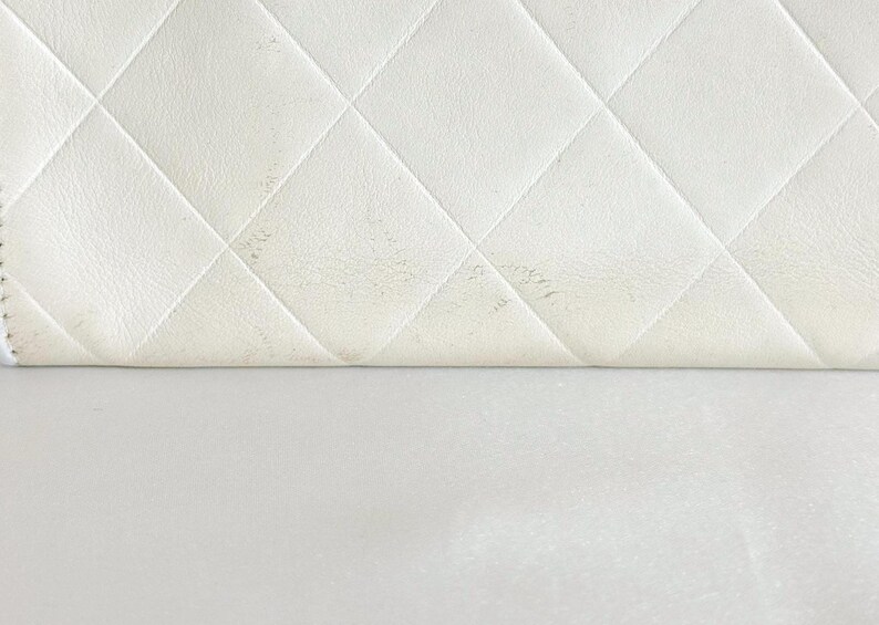 May include: Close-up of a white, quilted leather surface with a diamond pattern. The leather is slightly folded over, revealing a smooth, white surface below. Some areas of the leather show minor discoloration.