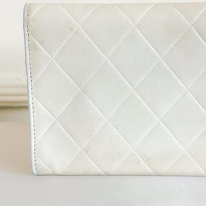 May include: Close-up of a white, quilted leather wallet. The wallet features a diamond pattern and a white border. The leather shows some signs of wear and discoloration. The wallet is on a white surface.