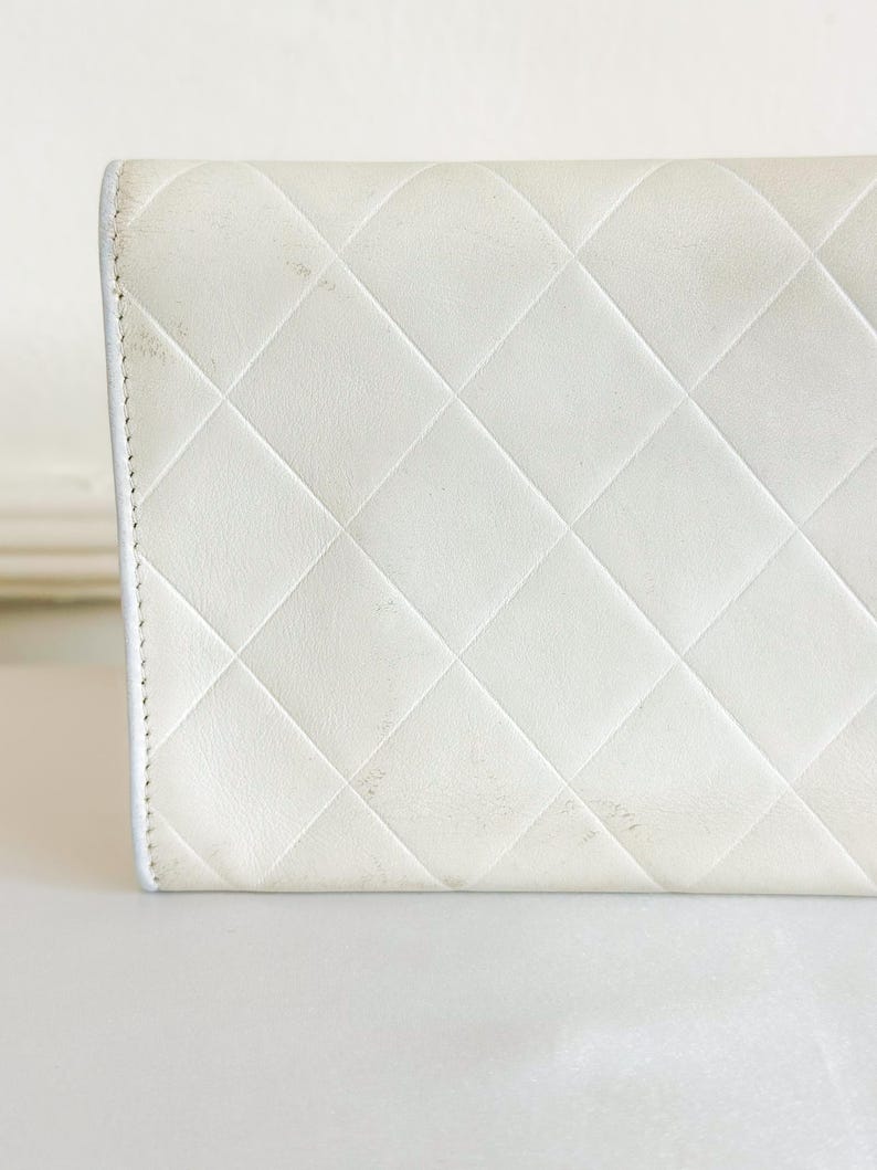 May include: Close-up of a white, quilted leather wallet. The wallet features a diamond pattern and a white border. The leather shows some signs of wear and discoloration. The wallet is on a white surface.