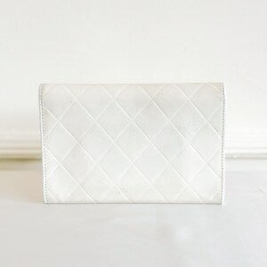 May include: A white, quilted leather wallet with a diamond pattern. The wallet is rectangular and has a simple, elegant design. The stitching is visible along the edges.