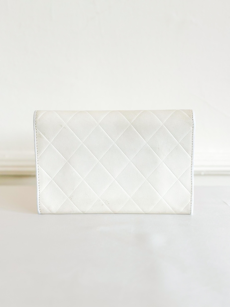 May include: A white, quilted leather wallet with a diamond pattern. The wallet is rectangular and has a simple, elegant design. The stitching is visible along the edges.