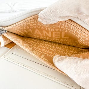 May include: Open white leather wallet with a silver zipper. The interior lining is tan and features the brand name "CHANEL" printed repeatedly. The wallet is held open, revealing the interior compartments.