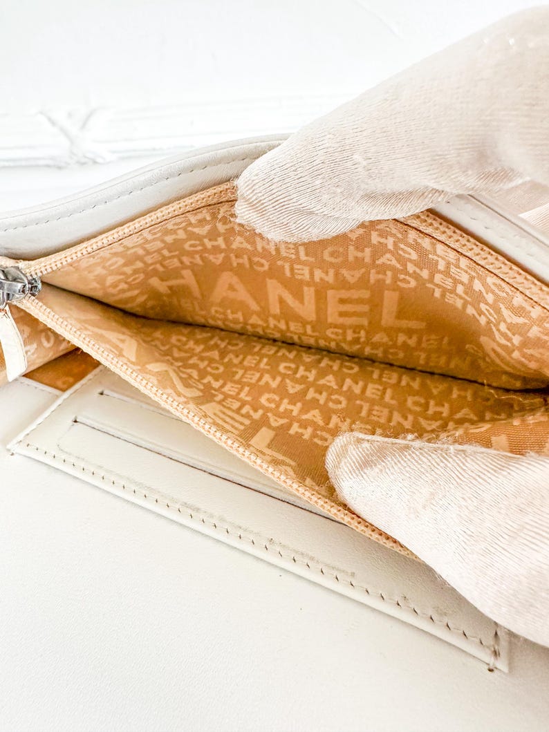 May include: Open white leather wallet with a silver zipper. The interior lining is tan and features the brand name "CHANEL" printed repeatedly. The wallet is held open, revealing the interior compartments.