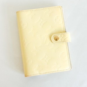 May include: Pale yellow leather organizer with embossed floral and logo pattern. Features a snap closure with a gold-tone button. The organizer has a glossy finish and a stitched border.