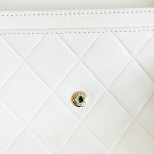 May include: Close-up of a white, quilted leather wallet. The wallet features a diamond pattern and a gold-colored button clasp with the brand name. The stitching is visible along the edges.