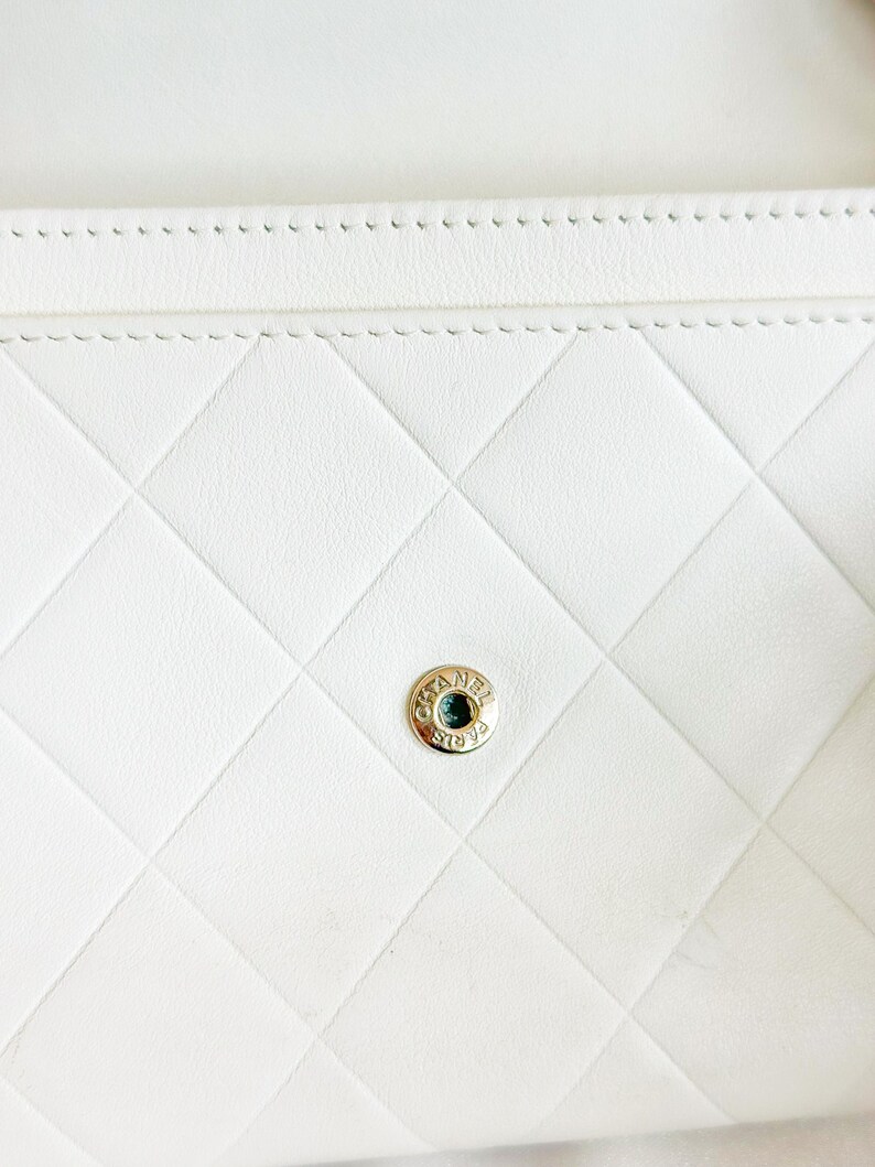 May include: Close-up of a white, quilted leather wallet. The wallet features a diamond pattern and a gold-colored button clasp with the brand name. The stitching is visible along the edges.