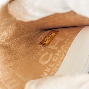 May include: Interior view of a cream-colored leather handbag. The lining is a light tan color with the embossed word "CHANEL" visible. A small, rectangular label with the number "18431555" is also visible.