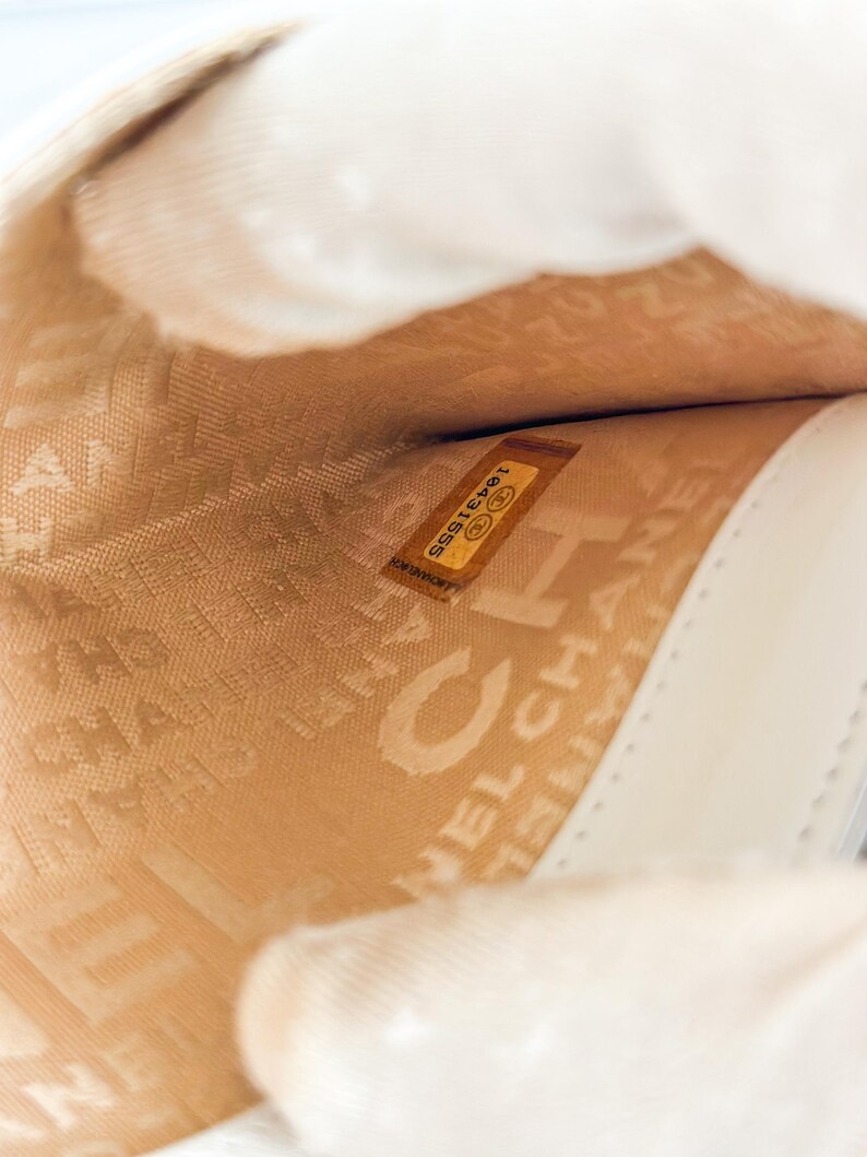 May include: Interior view of a cream-colored leather handbag. The lining is a light tan color with the embossed word "CHANEL" visible. A small, rectangular label with the number "18431555" is also visible.