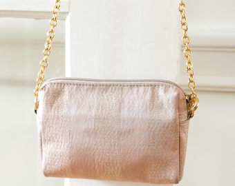 May include: A small, rectangular, blush-colored crossbody bag with a gold chain strap. The bag has a textured, metallic finish and a zipper closure. The chain is attached to the bag with small gold rings.