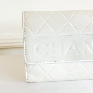 May include: Close-up of a white quilted leather wallet. The wallet features a diamond pattern and the embossed word "CHAN" across the front. The edges are neatly stitched, and the overall design is clean and elegant.