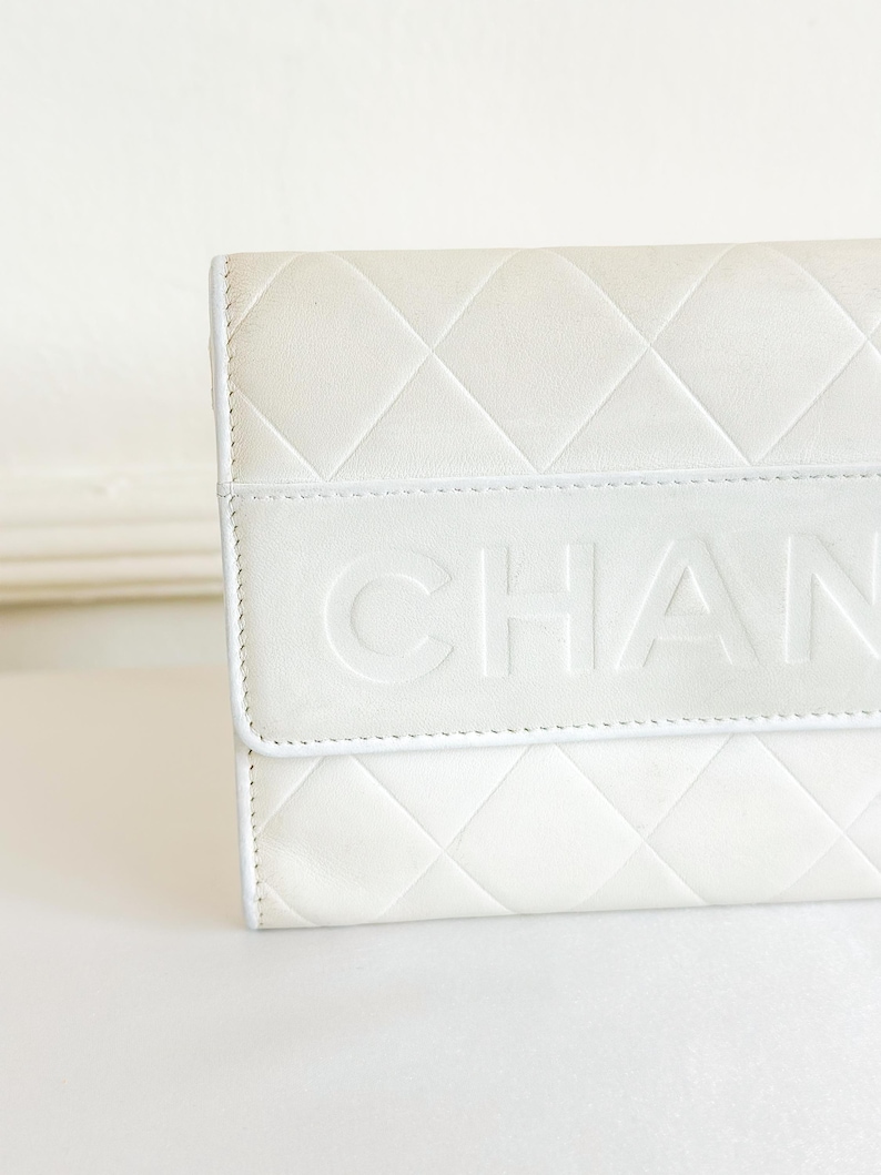 May include: Close-up of a white quilted leather wallet. The wallet features a diamond pattern and the embossed word "CHAN" across the front. The edges are neatly stitched, and the overall design is clean and elegant.