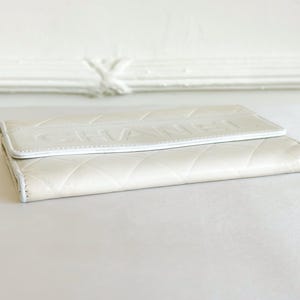 May include: Ivory-colored wallet with a quilted pattern and the embossed word "CHANEL" on the flap. The wallet is made of a soft, textured material and has a white trim.