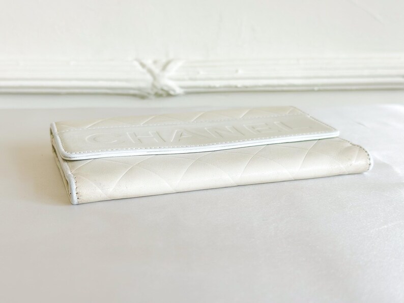 May include: Ivory-colored wallet with a quilted pattern and the embossed word "CHANEL" on the flap. The wallet is made of a soft, textured material and has a white trim.
