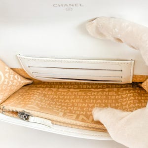 May include: White leather wallet with the CHANEL logo embossed on the top. The interior features a card slot and a zippered compartment. The lining is beige with the CHANEL logo printed repeatedly.
