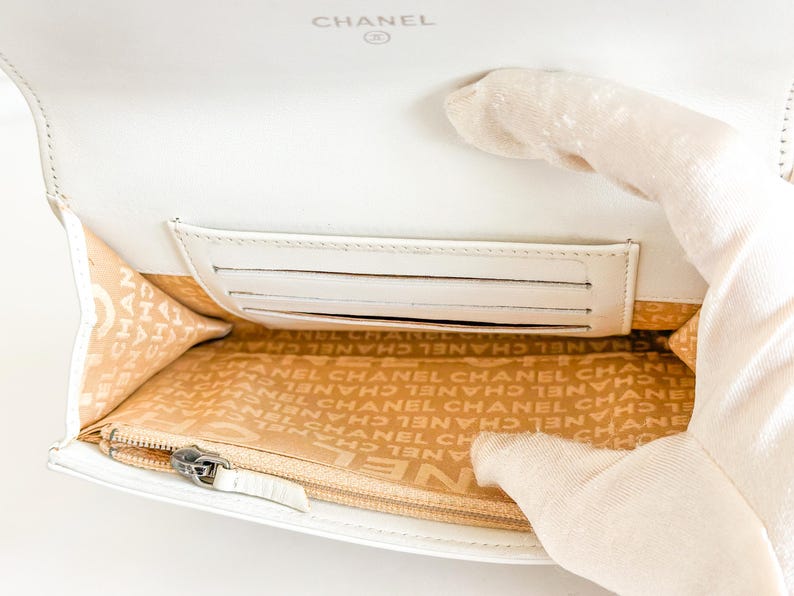 May include: White leather wallet with the CHANEL logo embossed on the top. The interior features a card slot and a zippered compartment. The lining is beige with the CHANEL logo printed repeatedly.