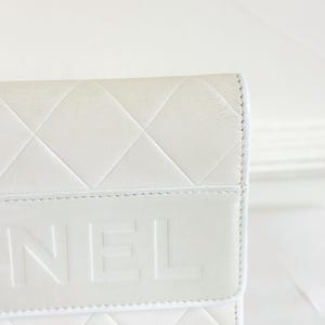 May include: Close-up of a white leather wallet with a quilted diamond pattern. The word "NEL" is embossed on the flap. The wallet has a clean, minimalist design.