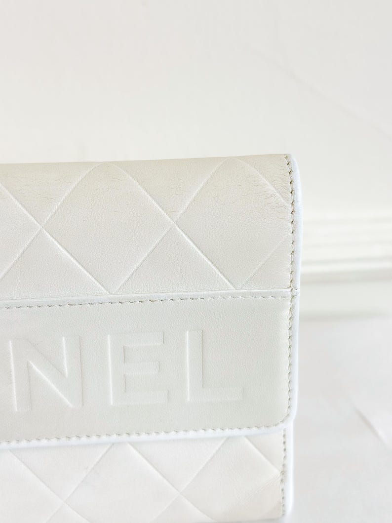 May include: Close-up of a white leather wallet with a quilted diamond pattern. The word "NEL" is embossed on the flap. The wallet has a clean, minimalist design.