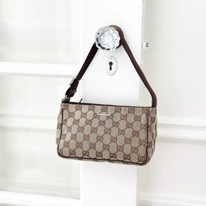 May include: A small, rectangular Gucci handbag with a brown and tan GG monogram pattern. The bag has a dark brown strap and a silver-tone buckle. The bag is hanging on a white door.