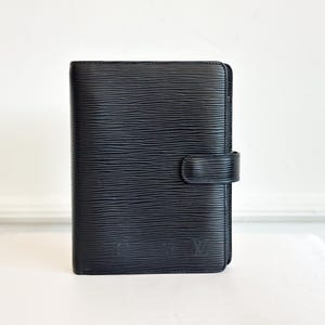 May include: Black leather organizer with a textured surface and a flap closure. The organizer has a small embossed logo on the lower right corner. The item is sitting on a white surface.