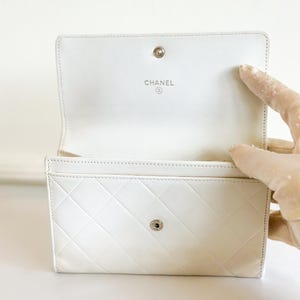 May include: Open white leather wallet with a diamond-stitched pattern. The interior features the brand name "CHANEL" in silver lettering. The wallet has a snap closure and is open, revealing card slots.