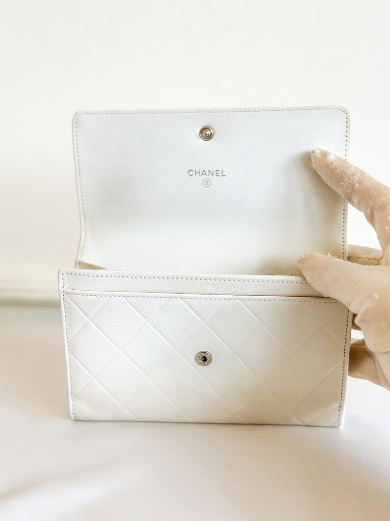 May include: Open white leather wallet with a diamond-stitched pattern. The interior features the brand name "CHANEL" in silver lettering. The wallet has a snap closure and is open, revealing card slots.